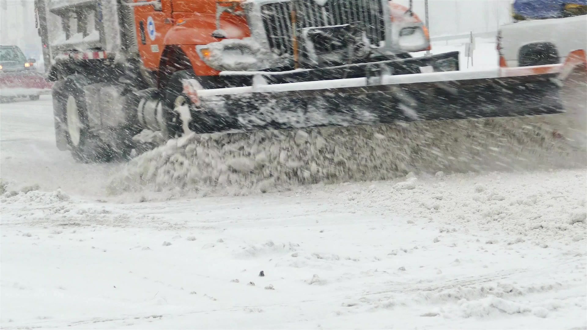 An orange plow truck pushing heavy snow off a road during a storm