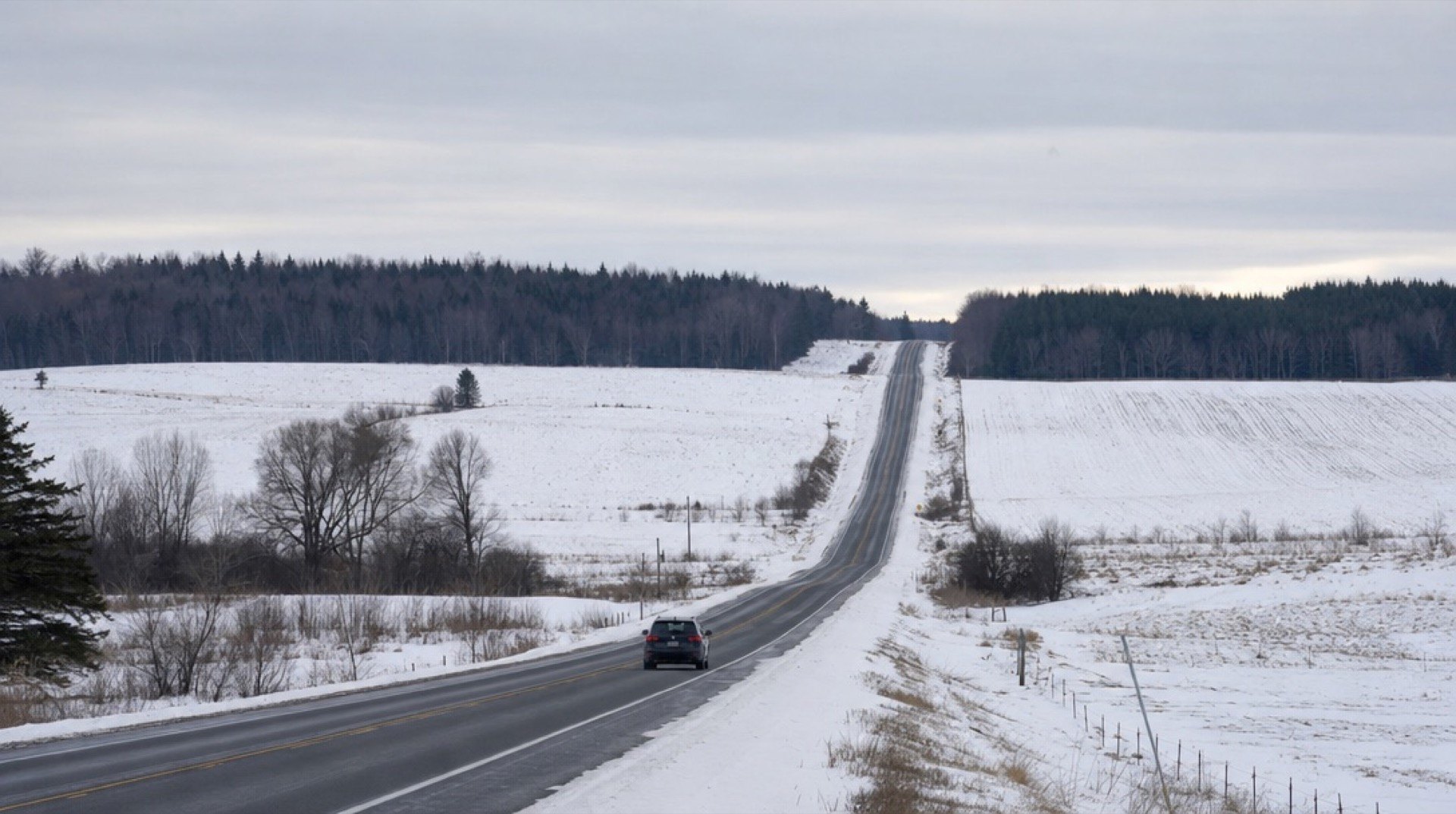 A single car on a long, snow-lined two-lane road through farmland in winter