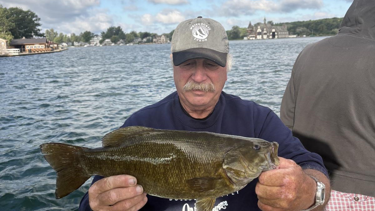 An angler on a boat holding a large smallmouth bass, with the Boldt Yacht House across the water