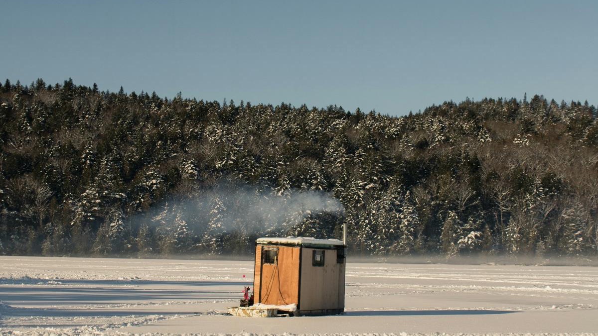 A small wooden ice shanty with smoke drifting from its stovepipe on a frozen bay below snowy hills
