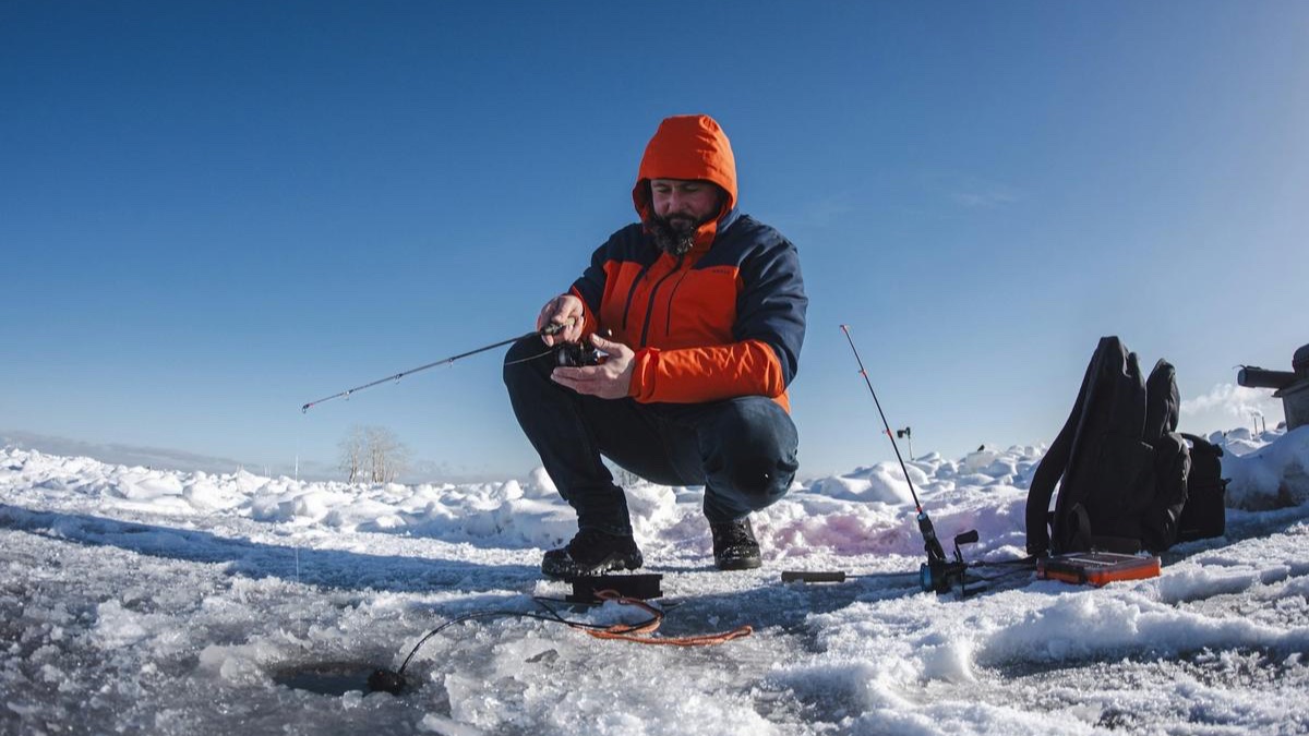 An angler crouched beside a hole in the ice, jigging with a short rod under a bright blue sky