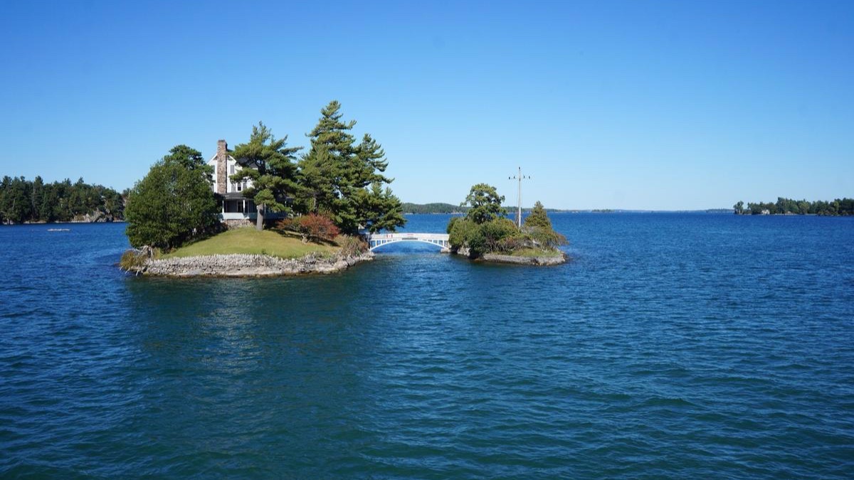A small island with a cottage and pine trees, joined to a tiny neighbor island by a footbridge, in deep blue river water